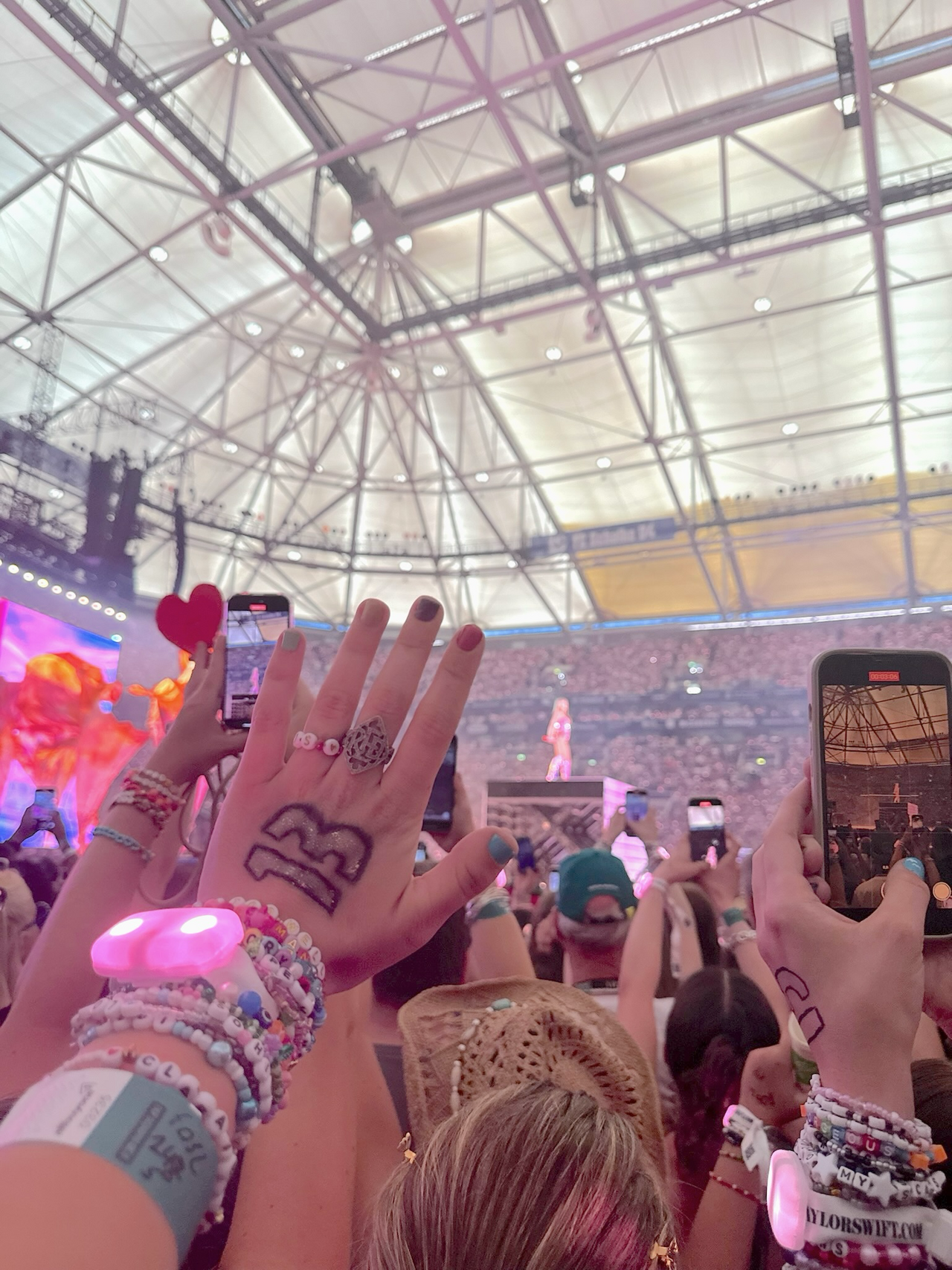 In the foreground, a fan raises their hand in the air with the number 13 written on it. The person is wearing friendship bracelets and a xyloband on their arm, glowing in pink. In the background, Taylor Swift can be seen in a pink outfit on a stage at The Eras Tour on July 17, 2024 in Gelsenkirchen. — Gelsenkirchen, July 17, 2024